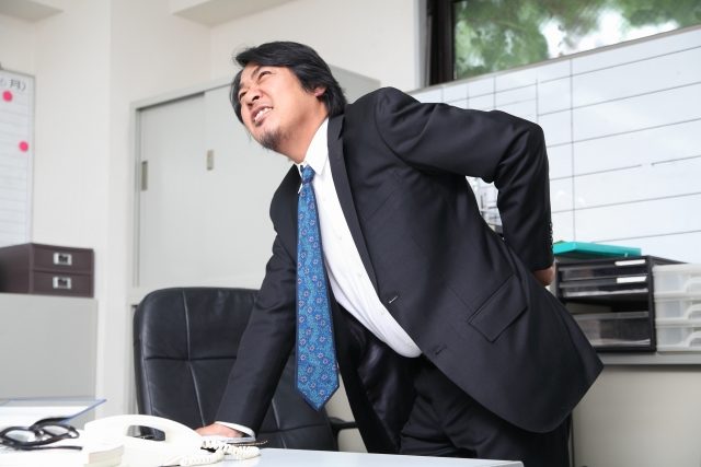 デスクワーク中に腰を押さえる男性のイメージ